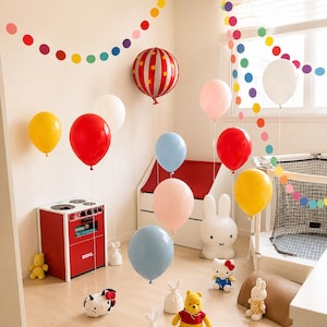 May include: A collection of colorful balloons in red, yellow, light blue, pink, and white, including one with a red and white striped pattern with yellow stars. A red Brio toy stove, a white rabbit lamp, and plush toys are arranged in the room.