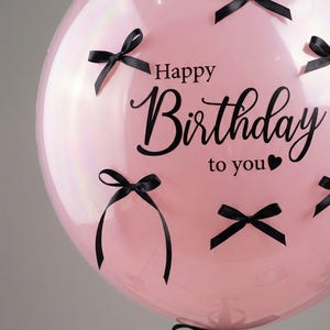 Puede incluir: Un globo rosa grande con la frase "Happy Birthday to you" en escritura elegante y lazos negros. El globo está decorado con varios lazos negros pequeños y un lazo más grande en la base.