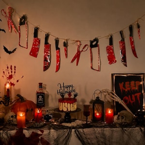 Puede incluir: Un conjunto de decoración de Halloween con una guirnalda de cuchillos y tijeras de papel manchados de sangre roja, un pastel con un adorno negro de Happy Halloween, un letrero de Keep Out, calaveras, velas naranjas y una botella Poison.