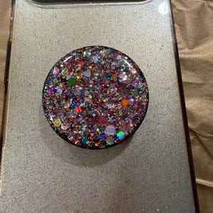 May include: A round phone grip with a black border, covered in colorful glitter. The glitter includes various shapes and colors, including pink, purple, green, and silver. The grip is attached to a silver phone case.