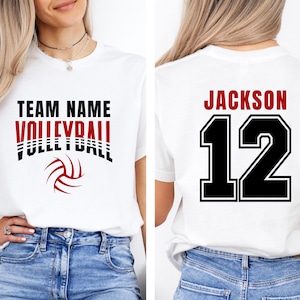 May include: White t-shirt with "TEAM NAME VOLLEYBALL" text in black and red, and a volleyball graphic. The back of the shirt displays "JACKSON 12" in red and black. The shirt is worn with blue jeans.