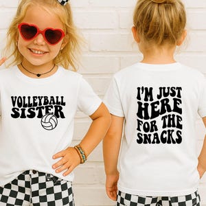 May include: White t-shirts with black text. One shirt says "Volleyball Sister" with a volleyball graphic. The other shirt says "I'm Just Here For The Snacks". The shirts are worn by a young child.