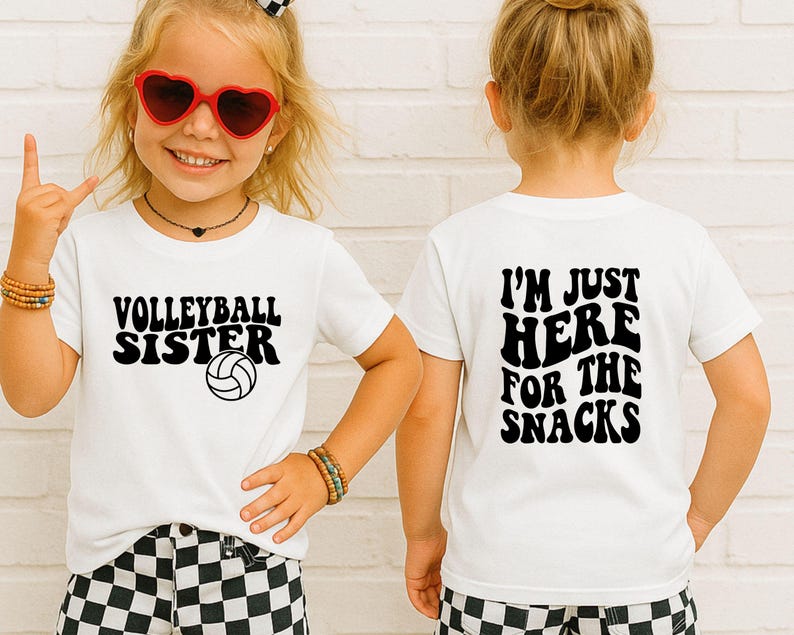 May include: White t-shirts with black text. One shirt says "Volleyball Sister" with a volleyball graphic. The other shirt says "I'm Just Here For The Snacks". The shirts are worn by a young child.