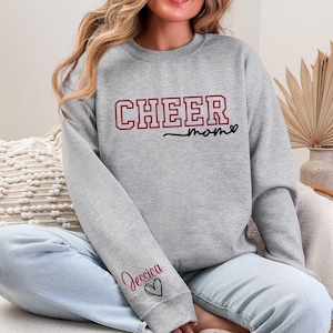 May include: A gray sweatshirt with the words "CHEER mom" in maroon and black lettering. The name "Jessica" is printed on the sleeve in maroon with a black heart. The sweatshirt is a casual garment.