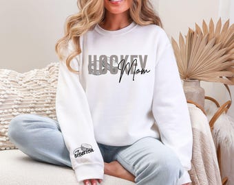 May include: White sweatshirt with the text "HOCKEY Mom" in gray and black script. A hockey puck graphic is incorporated into the word "HOCKEY". The sleeve features a hockey puck graphic with the name "Jessica" below it.