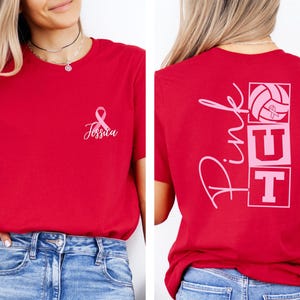 May include: Red t-shirt with a pink ribbon and the name "Jessica" printed on the front. The back of the shirt features the words "Pink Out" with a volleyball and the letters "UT" in pink. The shirt is made of a soft material.