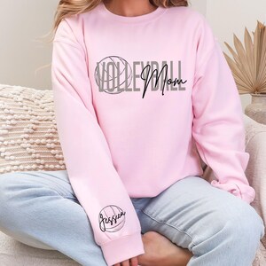 May include: A light pink sweatshirt with the words "Volleyball Mom" in gray and black. A volleyball graphic is above the word "Volleyball". The name "Jessica" is printed on the left sleeve, inside a volleyball graphic.