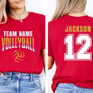 May include: Red volleyball t-shirt with the words "TEAM NAME VOLLEYBALL" in white and gold. A volleyball graphic is below the text. The back of the shirt has the name "JACKSON" and the number "12" in white with a gold outline.