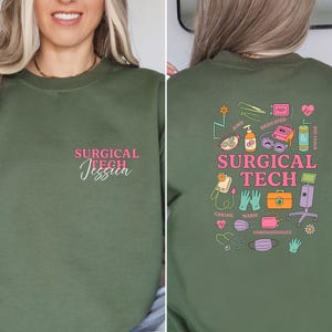 May include: An olive green sweatshirt with the text "SURGICAL TECH Jessica" in pink on the front. The back features a colorful graphic with medical-related icons and the words "SURGICAL TECH".