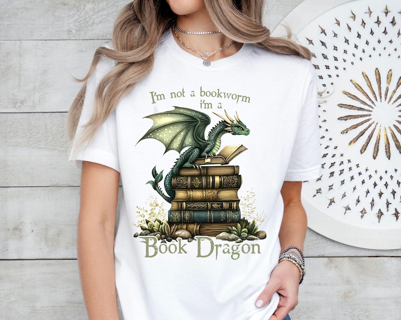 May include: White t-shirt with a green dragon sitting on a stack of books. The text on the shirt reads "I'm not a bookworm I'm a Book Dragon".
