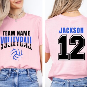 May include: Pink volleyball t-shirt with the words "TEAM NAME VOLLEYBALL" in black and blue. A blue volleyball graphic is below the text. The back of the shirt has the name "JACKSON" and the number "12" in blue and black.