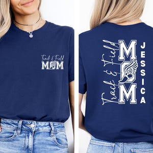 May include: Navy blue t-shirt with white text and graphics. The front of the shirt says "Track & Field MOM" with a winged shoe graphic. The back of the shirt says "Track & Field MOM" with a winged shoe graphic and the name "Jessica".