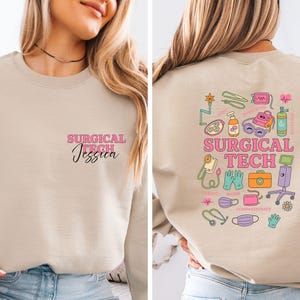 May include: A beige sweatshirt with the text "SURGICAL TECH Jessica" in pink and black on the front. The back features colorful medical-themed illustrations and the words "SURGICAL TECH".