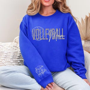 May include: Royal blue sweatshirt with the words "VOLLEYBALL Mom" in silver and white script. A volleyball graphic is integrated into the word "VOLLEYBALL". The sleeve features the name "Jessica Arthur" in a cursive font.