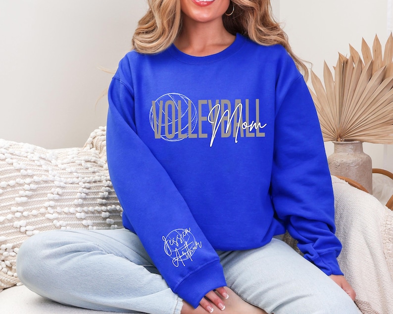 May include: Royal blue sweatshirt with the words "VOLLEYBALL Mom" in silver and white script. A volleyball graphic is integrated into the word "VOLLEYBALL". The sleeve features the name "Jessica Arthur" in a cursive font.