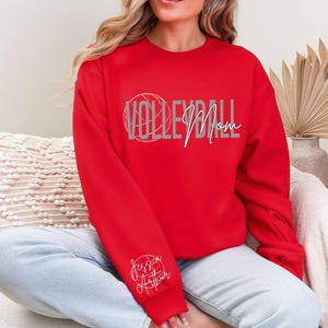 May include: A red sweatshirt with the words "VOLLEYBALL Mom" in a gray and white design. The word "Volleyball" is in a stylized font with a volleyball graphic. The name "Jessica Arthur" is written in cursive on the left sleeve.