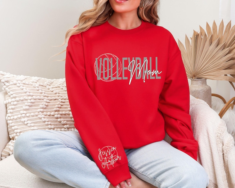 May include: A red sweatshirt with the words "VOLLEYBALL Mom" in a gray and white design. The word "Volleyball" is in a stylized font with a volleyball graphic. The name "Jessica Arthur" is written in cursive on the left sleeve.