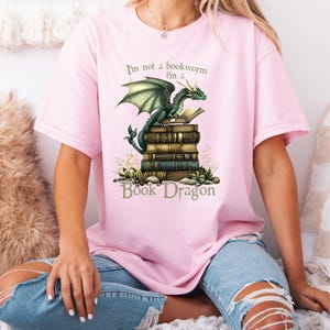 May include: Pink t-shirt with a green dragon perched on a stack of books. The text on the shirt reads "I'm not a bookworm I'm a Book Dragon".