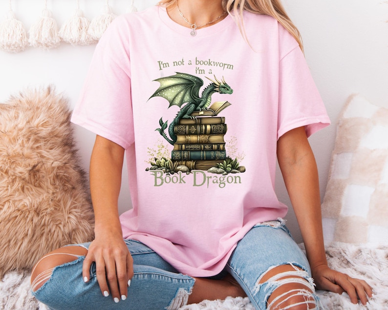 May include: Pink t-shirt with a green dragon perched on a stack of books. The text on the shirt reads "I'm not a bookworm I'm a Book Dragon".