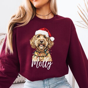 Pu&ograve; includere: Felpa bordeaux con un golden doodle con cappello da Babbo Natale e luci a stringa. Il nome del cane, Molly, &egrave; scritto in bianco sotto l'immagine. La felpa &egrave; di colore unito e ha maniche lunghe.