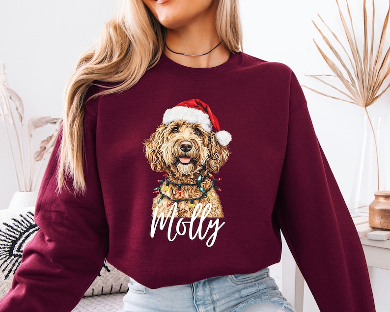 Pu&ograve; includere: Felpa bordeaux con un golden doodle con cappello da Babbo Natale e luci a stringa. Il nome del cane, Molly, &egrave; scritto in bianco sotto l'immagine. La felpa &egrave; di colore unito e ha maniche lunghe.