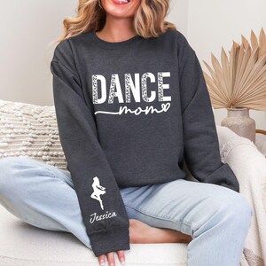 May include: A dark gray sweatshirt with the text "DANCE mom" in white with a leopard print pattern. The sweatshirt has a white silhouette of a dancer on the left sleeve with the name "Jessica" underneath.