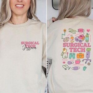 May include: Beige sweatshirt with the text "SURGICAL TECH Jessica" in pink and black on the front. The back features a colorful graphic with medical equipment and the words "SURGICAL TECH", and the words "Caring, Warm, Compassionate".