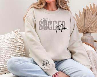 May include: Beige sweatshirt with the text "SOCCER Mom" in gray and black script. A soccer ball graphic is integrated into the word "SOCCER". The sleeve features a soccer ball graphic with the name "Mary" in script.