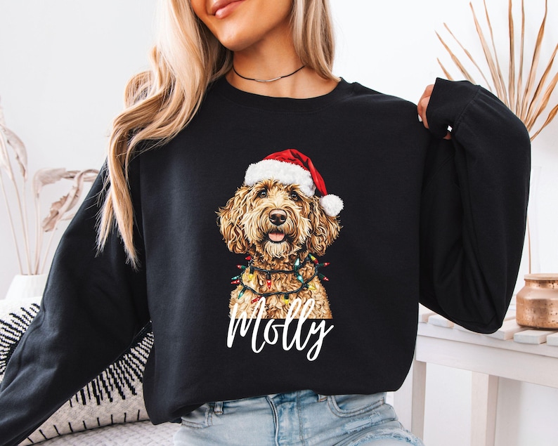 Pu&ograve; includere: Felpa nera con un cane golden doodle con cappello da Babbo Natale e luci a catena. Il nome del cane, "Molly", &egrave; scritto in bianco sotto. Un design festivo per le vacanze.
