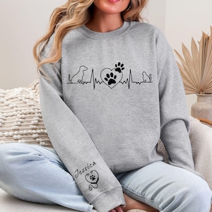 May include: A gray sweatshirt featuring a black graphic of a dog, a heart with paw prints, and a cat, all connected by a heartbeat line. The name "Jessica" is printed on the sleeve with a stethoscope and paw print design.