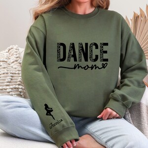 May include: Green sweatshirt with the text "Dance Mom" in black with a leopard print pattern. The sweatshirt has a small silhouette of a dancer on the left sleeve with the name "Jessica" underneath.
