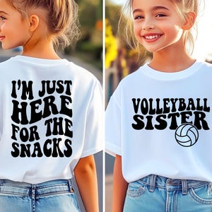 May include: Two white t-shirts with black text. One says "I'M JUST HERE FOR THE SNACKS" on the back. The other says "VOLLEYBALL SISTER" with a volleyball graphic. Both shirts are worn by a young person.
