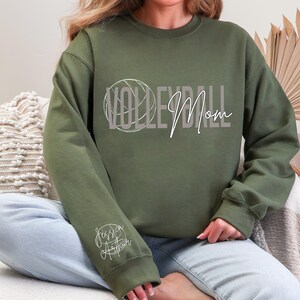 May include: An olive green sweatshirt with the words "VOLLEYBALL Mom" in a stylized font, with a volleyball graphic. The sleeve features the name "Jessica Arthur" in a cursive script. The sweatshirt is a casual, comfortable style.