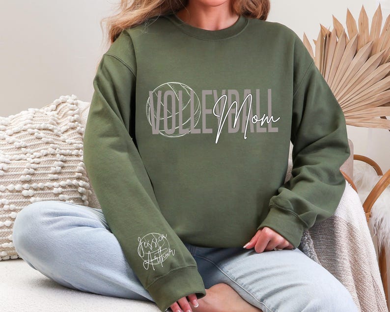 May include: An olive green sweatshirt with the words "VOLLEYBALL Mom" in a stylized font, with a volleyball graphic. The sleeve features the name "Jessica Arthur" in a cursive script. The sweatshirt is a casual, comfortable style.
