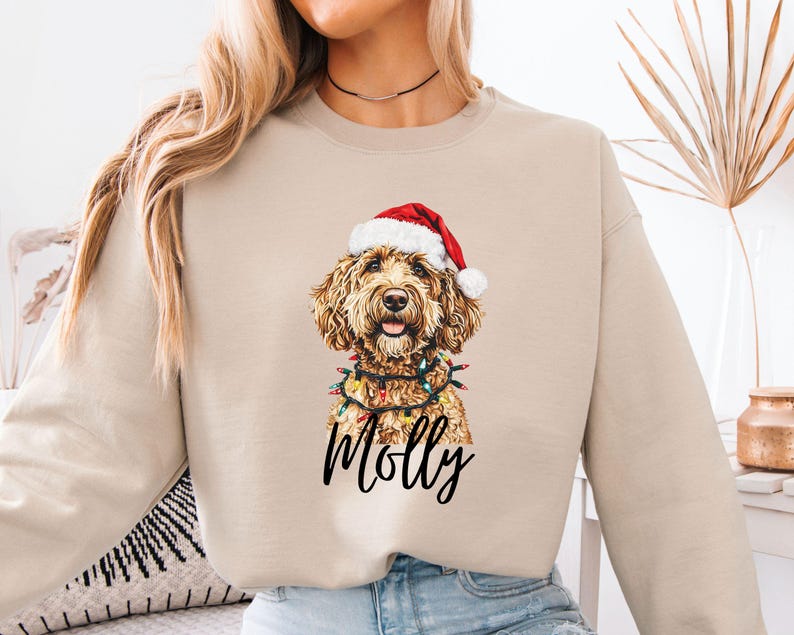 Pu&ograve; includere: Felpa beige con grafica di un cane con cappello di Babbo Natale e luci natalizie. Il nome del cane, "Molly", &egrave; scritto in nero sotto l'immagine. La felpa &egrave; di colore neutro, adatta all'abbigliamento casual.