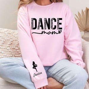 May include: Pink sweatshirt with the text "Dance Mom" in black with a leopard print design. The sweatshirt has a black silhouette of a dancer on the left sleeve with the name "Jessica" printed below.