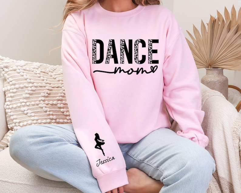 May include: Pink sweatshirt with the text "Dance Mom" in black with a leopard print design. The sweatshirt has a black silhouette of a dancer on the left sleeve with the name "Jessica" printed below.