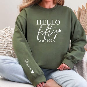 May include: An olive green sweatshirt with the words "HELLO fifty est. 1976" in white script. The sleeve has the name "Jessica" and a heart. The sweatshirt is a casual style.