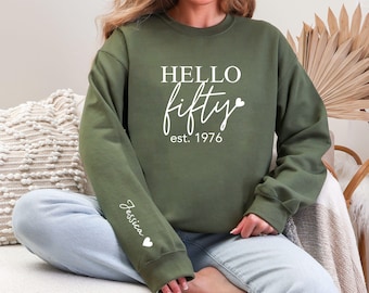 Può includere: Una felpa verde oliva con la scritta bianca "HELLO fifty est. 1976". La manica ha il nome "Jessica" e un cuore. La felpa è in stile casual.