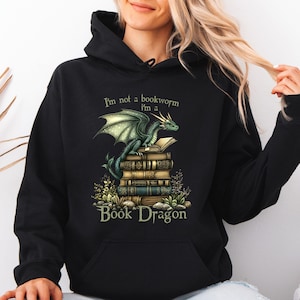 May include: Black hooded sweatshirt featuring a green dragon perched atop a stack of books. The text "I'm not a bookworm I'm a Book Dragon" is printed above and below the dragon illustration. The design includes details of the dragon's wings and the books' aged appearance.