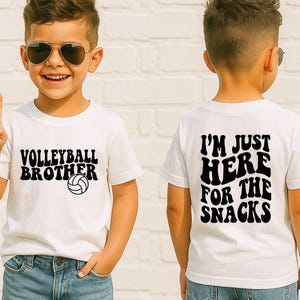 May include: A white t-shirt with the text "VOLLEYBALL BROTHER" and a volleyball graphic in black. The back of the shirt says "I'M JUST HERE FOR THE SNACKS" in black. The shirt is worn by a child.