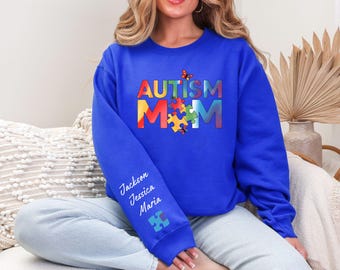 May include: Royal blue sweatshirt with the words "AUTISM MOM" in colorful letters and puzzle piece graphics. The sleeve features the names "Jackson Jessica Maria" in white script. A butterfly graphic is also present.