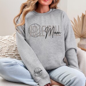 May include: A gray sweatshirt with the words "Volleyball Mom" in black script and a volleyball graphic. The sleeve features a volleyball graphic with the name "Jessica" in script. The sweatshirt is a casual, comfortable style.