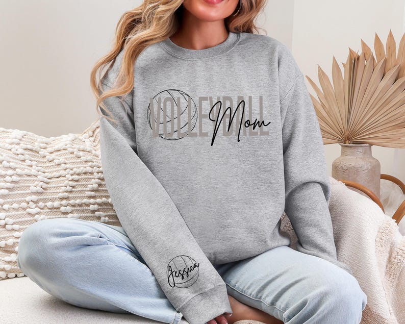 May include: A gray sweatshirt with the words "Volleyball Mom" in black script and a volleyball graphic. The sleeve features a volleyball graphic with the name "Jessica" in script. The sweatshirt is a casual, comfortable style.