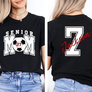 May include: A black t-shirt featuring a front graphic with the text SENIOR MOM and a soccer ball with the number 27 inside. The back of the shirt displays a large white number 7 with the name Jackson written in red script across the number.