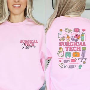 May include: A light pink sweatshirt with the text "SURGICAL TECH Jessica" on the front. The back of the sweatshirt features a colorful graphic with medical-related illustrations and the words "SURGICAL TECH".