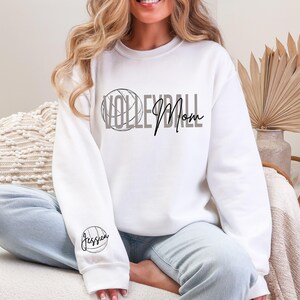 May include: White sweatshirt with the words "VOLLEYBALL Mom" in gray and black script. A volleyball graphic is incorporated into the word "VOLLEYBALL". The sleeve features the name "Jessica" with a volleyball graphic.