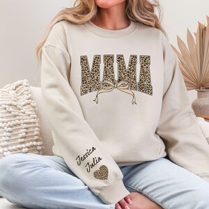 May include: Beige sweatshirt with the word "MAMA" in a leopard print design, with a bow detail. The sleeve features the names "Jessica Julia" and a leopard print heart. A cozy and stylish top.