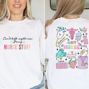 May include: White sweatshirt with a pink, blue, and green graphic of medical tools and the word "NURSE". The text on the front of the sweatshirt reads "Can't talk right now Doing NURSE STUFF".