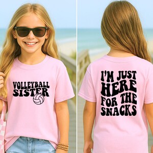 May include: A light pink t-shirt with the words "VOLLEYBALL SISTER" and a volleyball graphic. The back of the shirt says "I'M JUST HERE FOR THE SNACKS" in black text. The shirt is worn by a person with sunglasses.